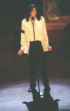 a woman standing in front of a microphone with her hands on her hips, wearing black pants and a white jacket