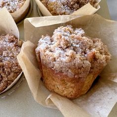 four muffins with powdered sugar on top are sitting in brown paper wrappers