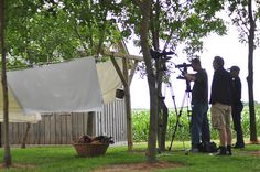 two men are standing in the grass near some trees and one is filming another man