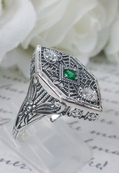 "Green Emerald & White Cubic Zirconia (CZ) Ring Charlotte Design#231 Made To Order Resurrected from the Art Deco movement, this gorgeous Art Deco reproduction filigree ring is crafted in sterling silver. This ring is marked 925 for solid sterling. This lovely filigree ring is set with a stunning quality 2mm center round-cut simulated emerald gemstone. Two small white cubic zirconia (CZ) gemstones also grace the face of the ring, one near each point. There are 2 filigree accent flowers that g Art Deco Emerald Ring With Diamond Accents As Gift, Silver Diamond Ring With May Birthstone Accents, Vintage Silver Emerald Ring With Diamond Accents, Silver Emerald Ring With Diamond Accents Art Deco, Art Deco Silver Emerald Ring With Diamond Accents, Filigree May Birthstone Ring, May Birthstone Filigree Rings, Antique Cubic Zirconia Ring For Gift, Victorian Emerald Ring With Diamond For Gift