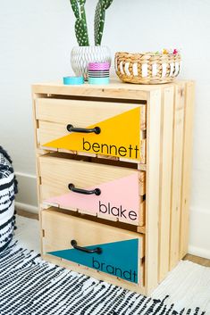 a wooden dresser with three drawers and colorful labels on the bottom drawer, next to a potted plant