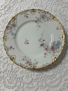 a white and gold plate sitting on top of a doily covered table with pink flowers