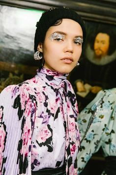 a woman with silver eyeshades and flowers on her dress is standing in front of a painting