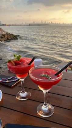 two cocktails sitting on top of a wooden table next to the ocean