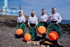 three women in white shirts are holding orange and green baskets on the beach near water