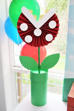 a paper flower in a vase with balloons and a clock on the window sill