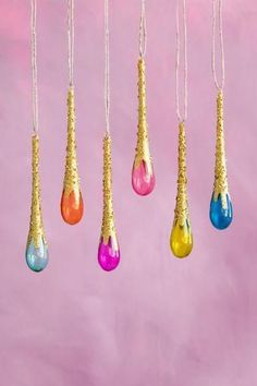 five different colored glass tears hanging from gold chains on a pink background with white string