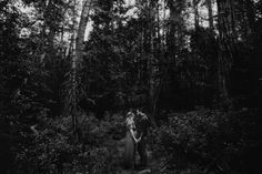 black and white photo of two people standing in the woods with tall trees behind them