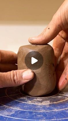 two hands are working with clay on a blue table top and another hand is holding the block