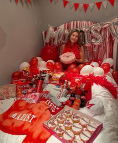 a woman sitting on a bed surrounded by red and white balloons, confetti, candy bars, and other items