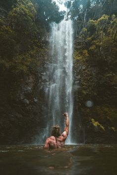 a man standing in the water next to a waterfall and reaching up into the air