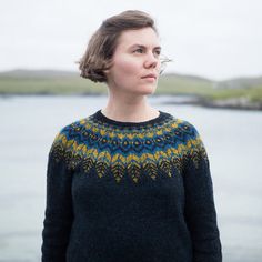 a woman standing in front of the water wearing a sweater with an intricate design on it