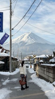 a woman walking down a snow covered street with a mountain in the background and power lines above her