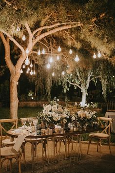 an outdoor dinner table set up under a tree at night with hanging lights and flowers