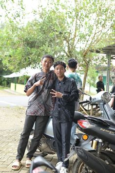 two young men standing next to each other near parked motor scooters and trees