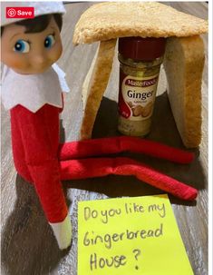 an elf is sitting in front of a gingerbread house with a sign that says do you like my gingerbread house?