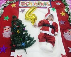 a baby dressed as santa claus laying on a blanket next to a christmas tree and balloon