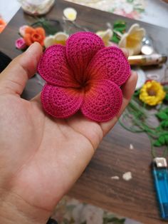 a hand holding a pink crocheted flower on top of a wooden table next to flowers