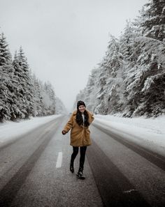 a woman walking down the middle of a road in front of some snow covered trees