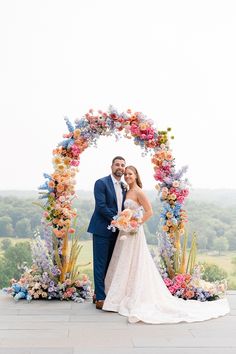 a bride and groom standing in front of an arch with colorful flowers on it at their wedding