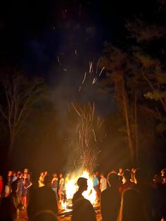 people gathered around a fire pit at night with fireworks in the sky and trees behind them