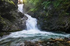 a waterfall in the middle of a forest with rocks and green plants on either side