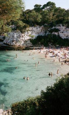 many people are swimming in the clear blue water on a beach with cliffs behind them