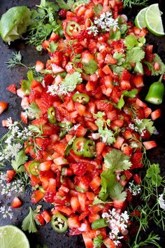 fresh strawberries and limes are arranged on a black surface with flowers, cilantro, and herbs