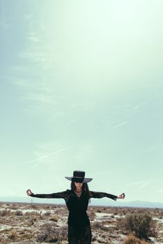 a woman in a black dress and hat standing on top of a dirt field with her arms outstretched