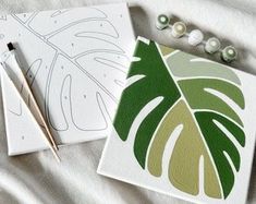 two cards with green leaves on them next to some knitting needles and beads, along with one card that has a leaf drawn on it