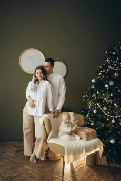 a man and woman standing next to a christmas tree in front of a baby on a chair