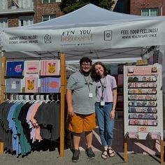 two people standing in front of a tent selling t - shirts and other items for sale