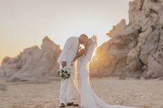 a bride and groom kissing on the beach in front of large rock formations at sunset