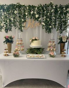 a white table topped with lots of cakes and desserts next to greenery covered walls