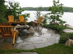 two wooden chairs sitting on top of a stone patio next to a body of water