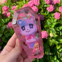 someone holding up a pink bear shaped object in front of some bushes and flowers with glitter on it