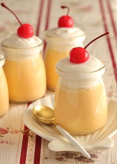 three small jars filled with pudding and topped with cherries on a white plate next to two spoons