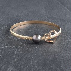 Price is per bangle. Multiple bangles are sold separately.  4mm Hawaiian Heirloom style bangle bracelet with a free moving Tahitian Pearl and a matching hand bent and soldered heart charm.  Two shape of Tahitian pearl available: 1. Tahitian pearl is round in shape, approximately 10-10.5mm in size. Some minor dimples/blemishes may be visible on the back or side of pearl. We choose a clean face for the front of the pearl facing outwards from the bangle.  2. Oval shape Tahitian pearl with or withou Pearl Bangles Gold, Hawaiian Heirloom Jewelry, Tahitian Pearls Jewelry, Heart Bangle Bracelet, Hammered Bangles, Stackable Bangles, Hawaiian Jewelry, Pearl Bangle, June Birthstone Jewelry