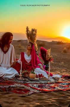 two women sitting on top of a blanket in the middle of a desert at sunset