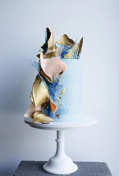 a blue cake with gold leaf decorations on it's top and bottom layer, sitting on a white pedestal