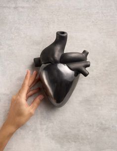 a hand holding a black heart sculpture on top of a cement floor next to a gray wall