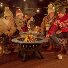 four people sitting around an outdoor fire pit with lights strung above them and one person holding a cup