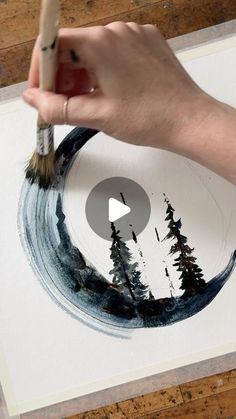 someone is using a brush to paint a painting on paper with trees in the background