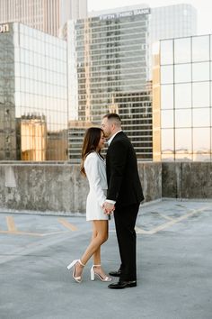 a man and woman standing next to each other in an empty parking lot with tall buildings behind them