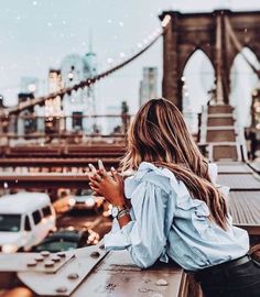 a woman sitting on top of a bench next to a bridge and looking at her cell phone