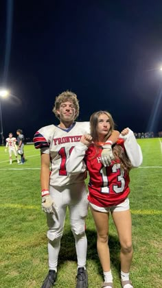 two people in football uniforms posing for a photo on the field at night with their arms around each other