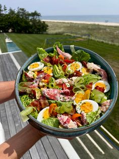 a person holding a bowl filled with salad on top of a wooden deck next to the ocean