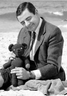 a man sitting on the beach with a teddy bear in his lap and wearing a suit