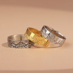 two wedding rings with mountains on them sitting on a table next to another ring that is gold and silver