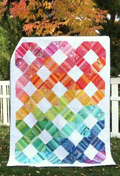 a colorful quilt hanging on the side of a white fence with trees in the background